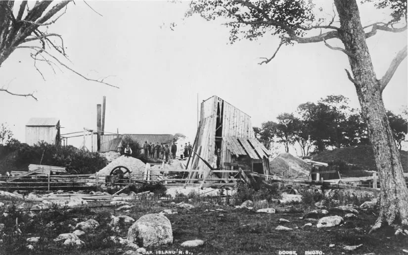 Black-and-white photograph of a wooden derrick and shed at the dig site, a line of men standing on the platform.