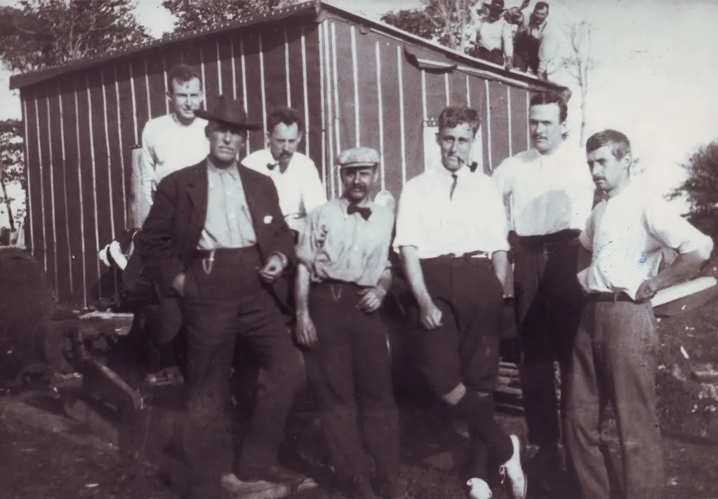 Eight men in shirtsleeves and hats posed in front of a corrugated metal shed at the dig camp.