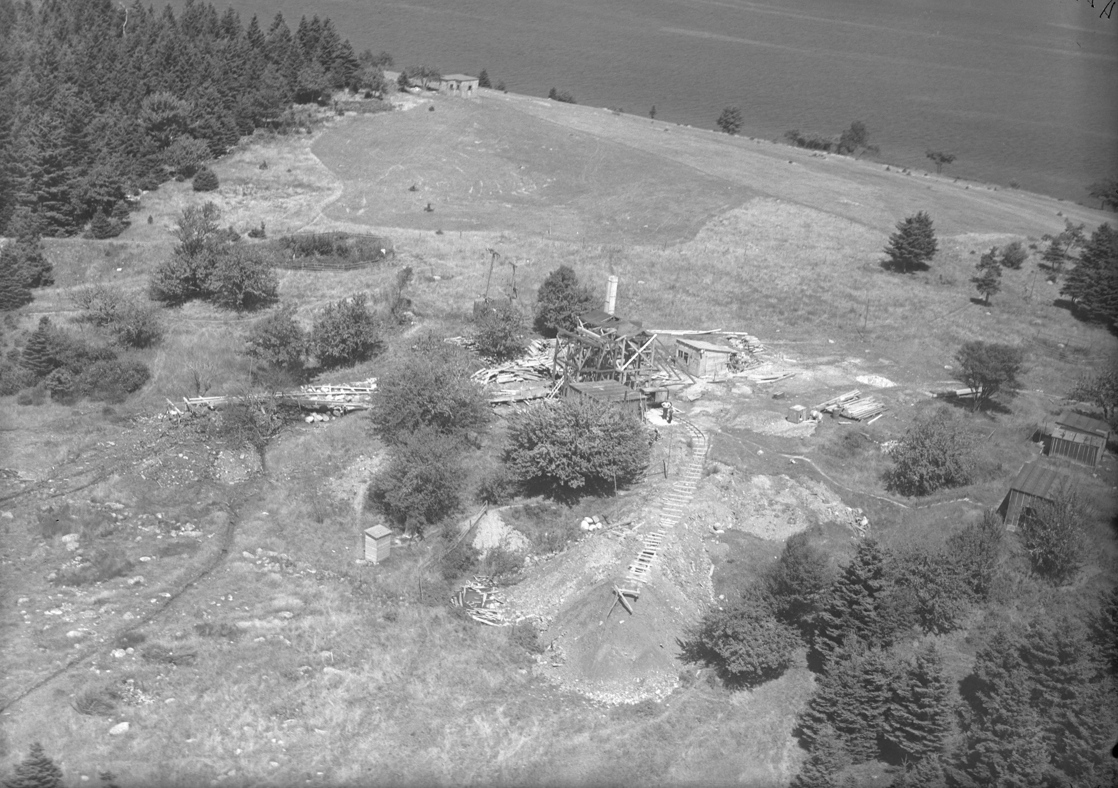 Aerial photograph of a cleared headland with a tall drilling rig, scattered buildings and spoil heaps among the trees.