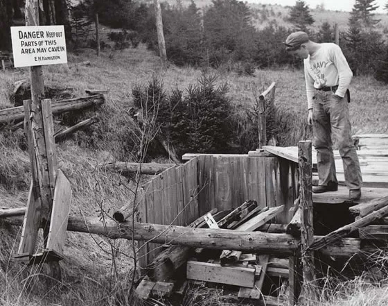A man in a cap standing at the edge of a cribbed shaft beside a hand-lettered sign warning that the ground may cave in.