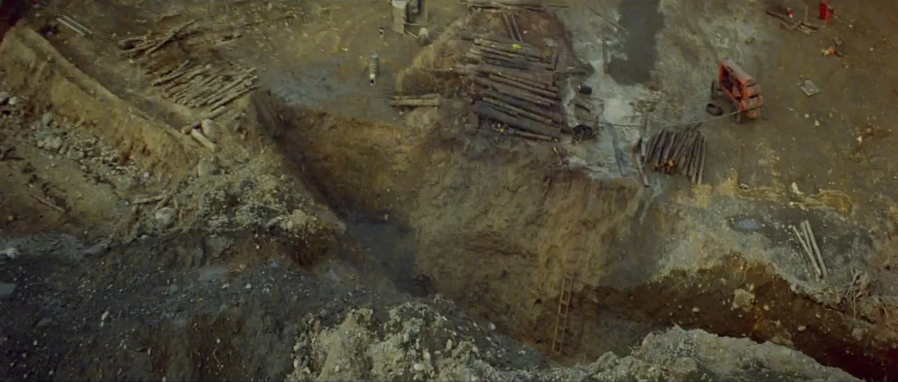 The same shoreline after the blast: a long trench cut through mud, timbers exposed, a red machine at the edge.