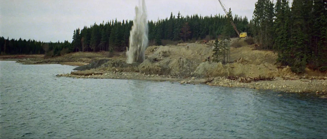 A plume of water and debris erupting from the shoreline as a charge goes off, seen from the water, with a crane on the bank.