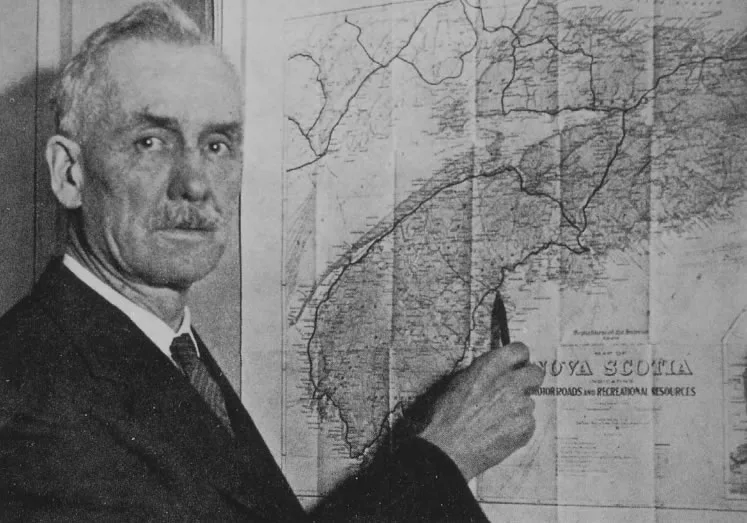 An older man in a suit and tie pointing a pen at a large wall map of Nova Scotia.