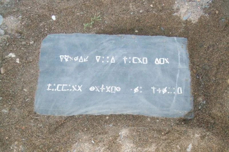 A flat dark slab lying on gravel, carved with two rows of angular symbols.