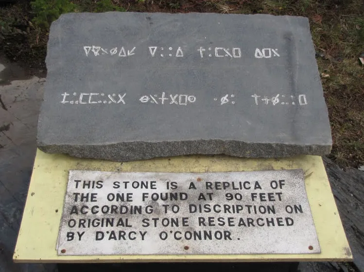 A flat grey stone slab carved with two rows of angular symbols, resting above an engraved metal plaque.
