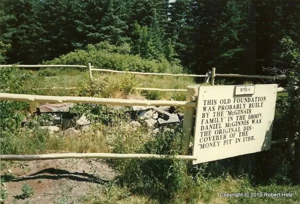 A low stone foundation in a grassy clearing behind a wooden rail fence, with a painted sign describing it.