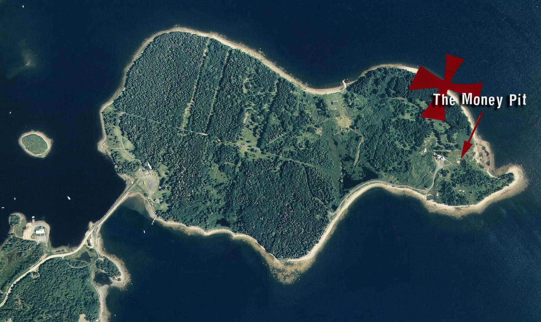 Aerial view of a wooded island in dark blue water, joined to the mainland by a narrow causeway; a red cross and arrow mark the Money Pit near the island's east end.